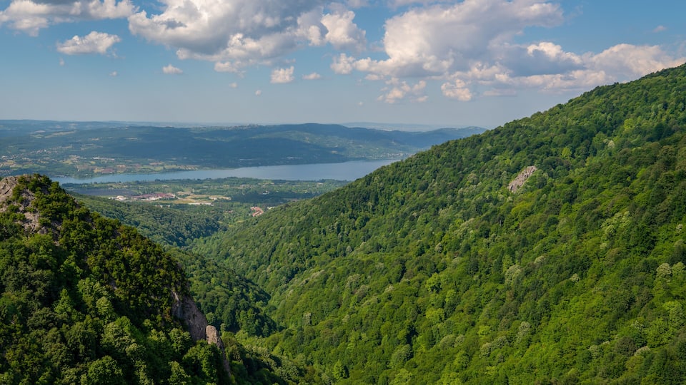 A mountainous landscape with dense green forests, a lake, and distant hills under a partly cloudy sky.