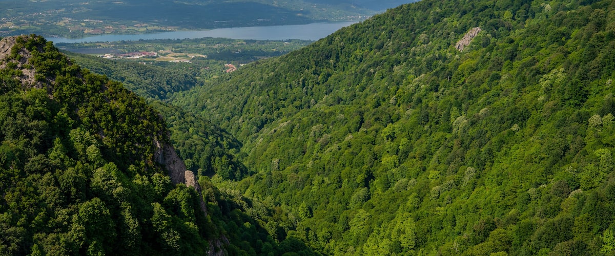 A mountainous landscape with dense green forests, a lake, and distant hills under a partly cloudy sky.