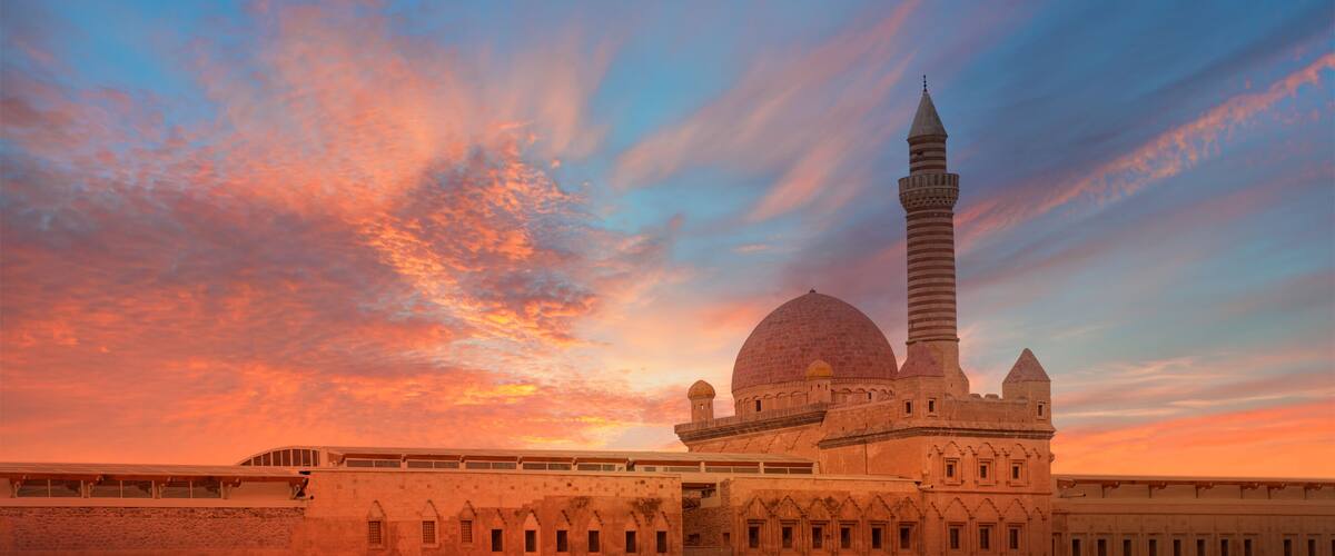 A mosque with a large dome and a tall minaret, set against a sunset sky.
