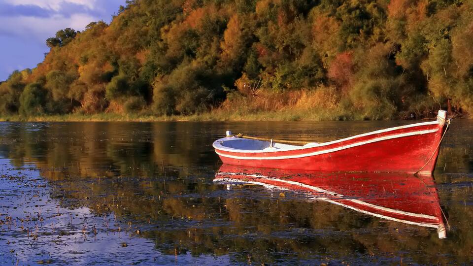 A red boat with white stripes on calm water, surrounded by autumn-colored trees.