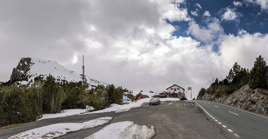 Uma estrada coberta de neve com uma montanha ao fundo, algumas casas e céu claro.