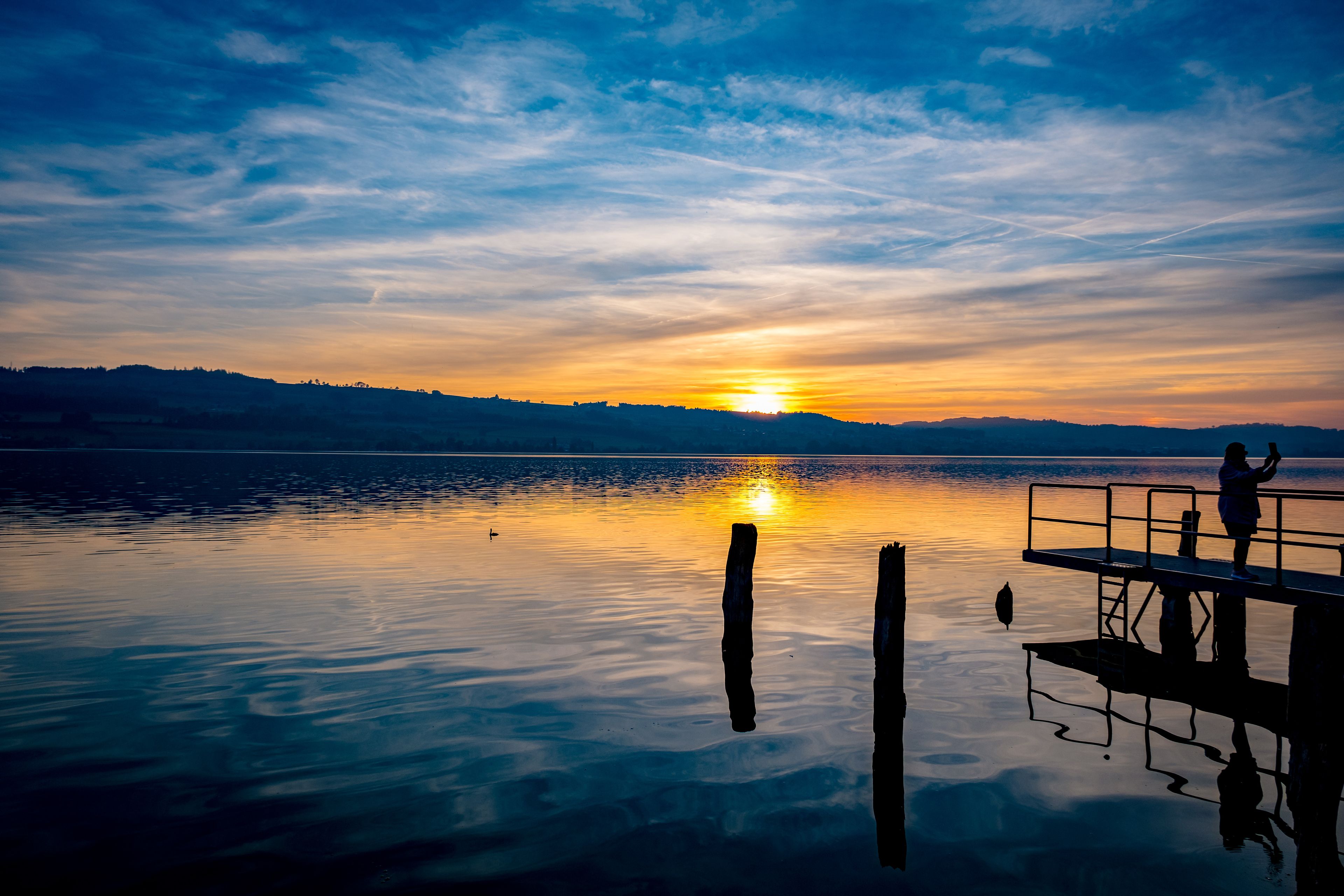 Un atardecer sobre un lago tranquilo con un muelle y una persona al final.