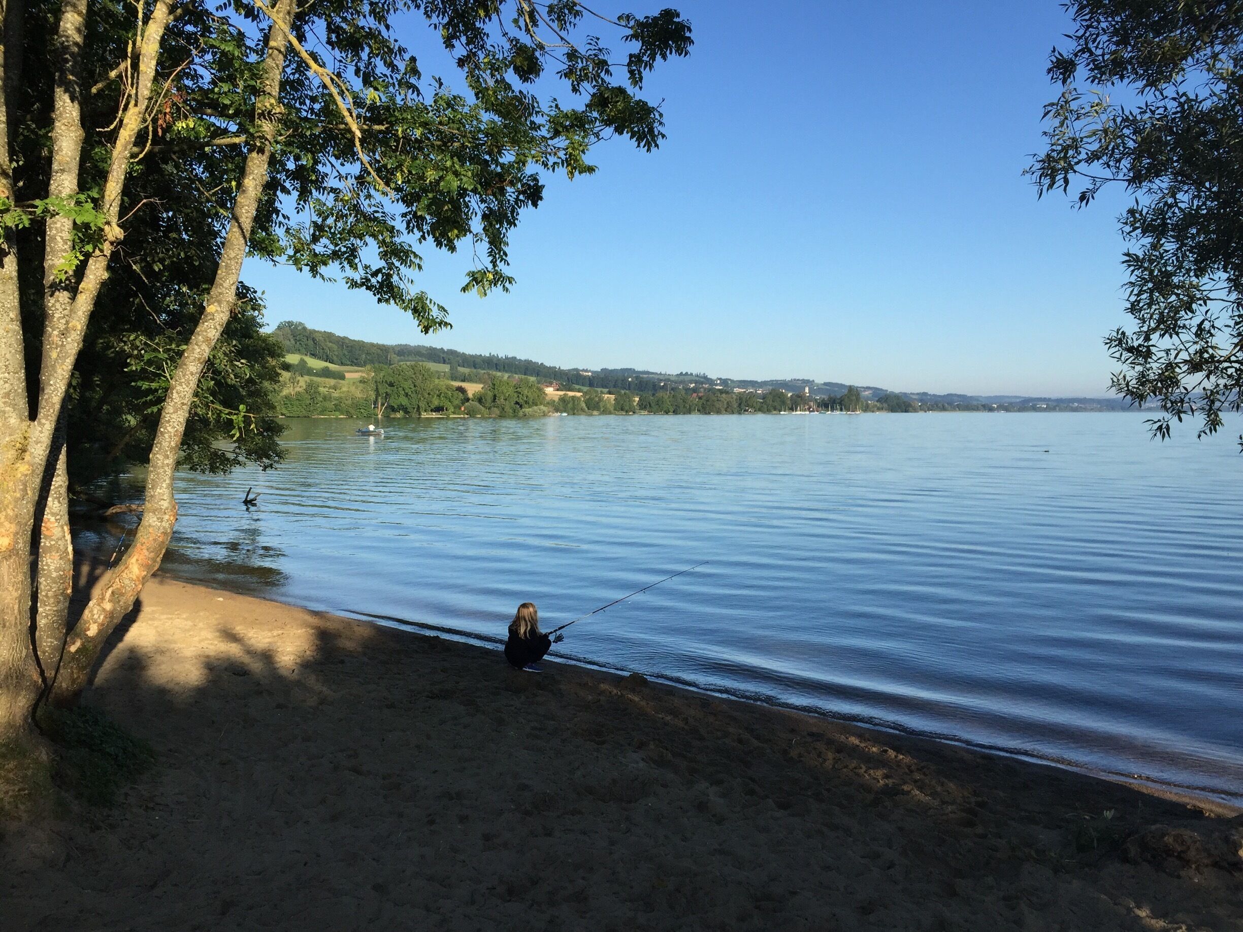 A calm lake with a person fishing, surrounded by trees and distant hills.