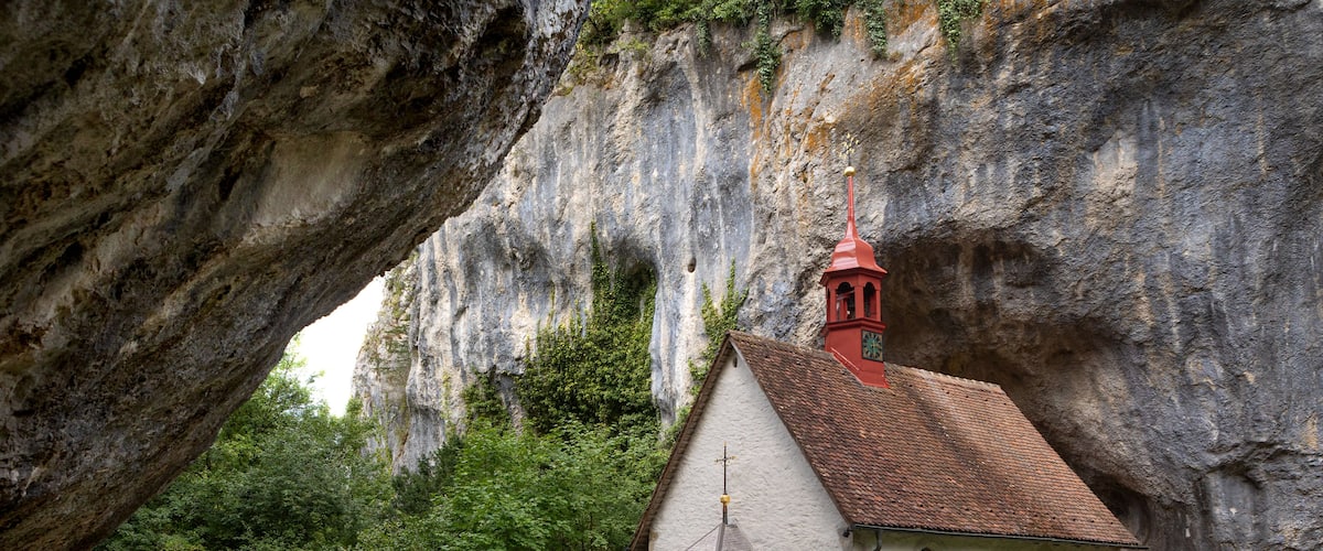 A small church with a red roof nestled in a rocky gorge, surrounded by lush greenery.