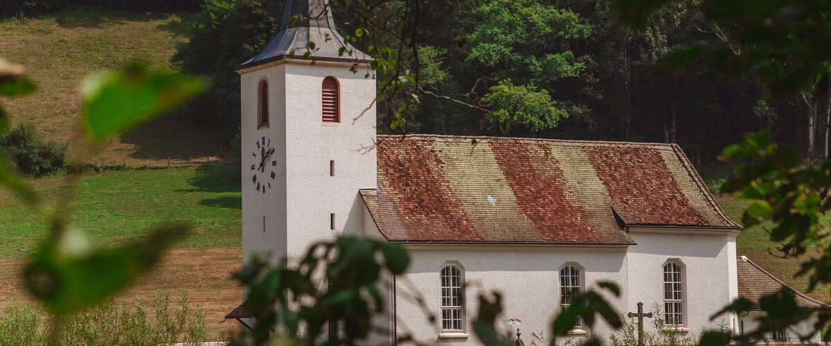 Une église dotée d’un clocher et d’un toit en tuiles, entourée d’arbres et d’un champ verdoyant.