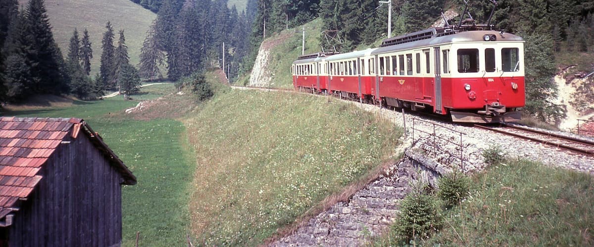 A red and white train traveling through a forested area with a small building on the left.