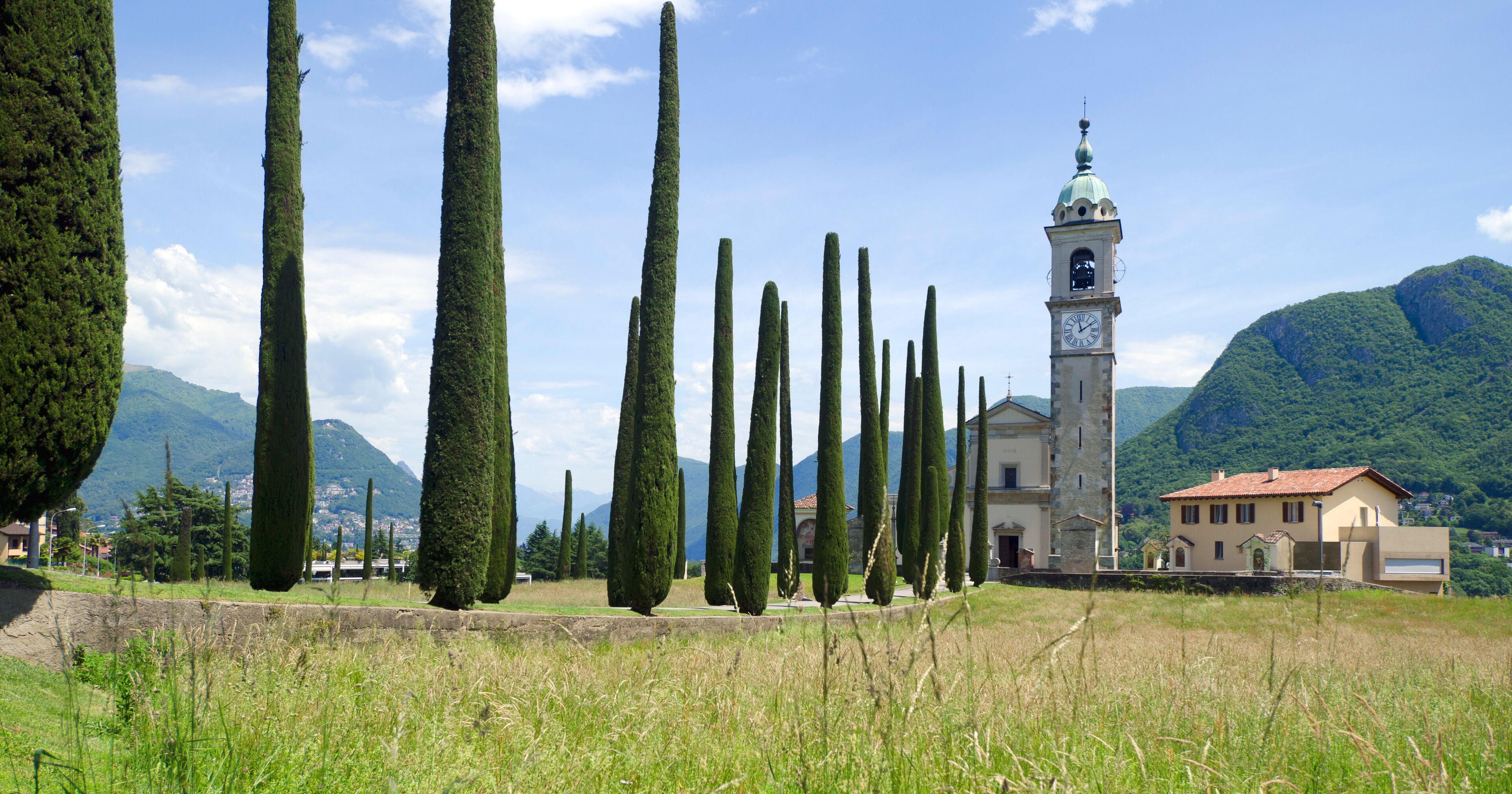 A tower with a clock, surrounded by tall cypress trees, set against a mountainous backdrop.