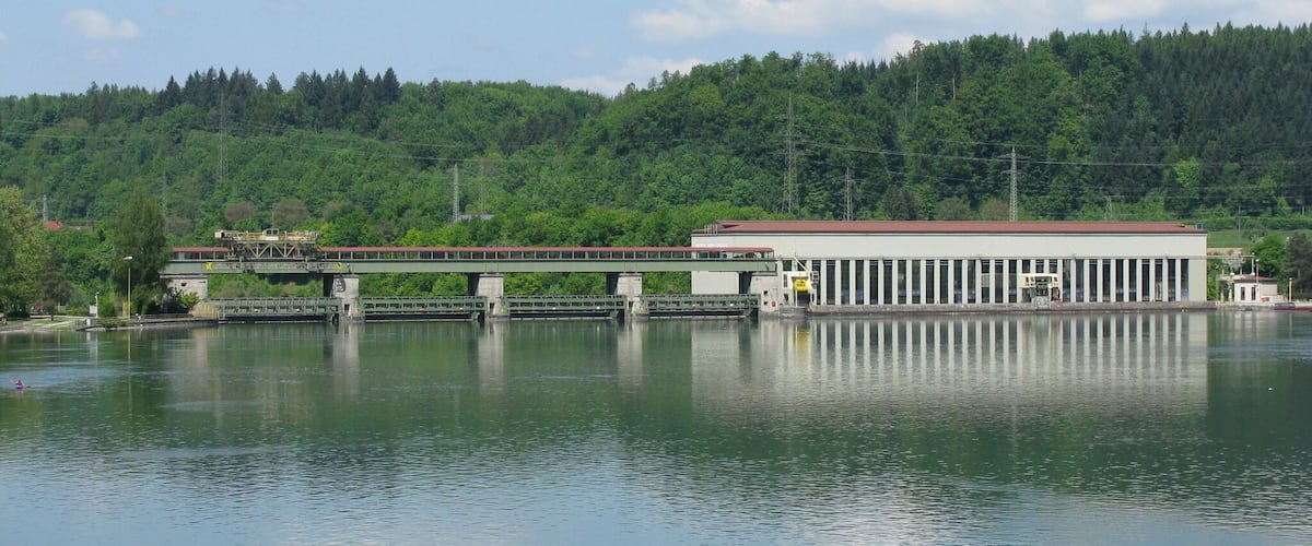 A bridge over a calm river with a building on the right and a forested hill in the background.