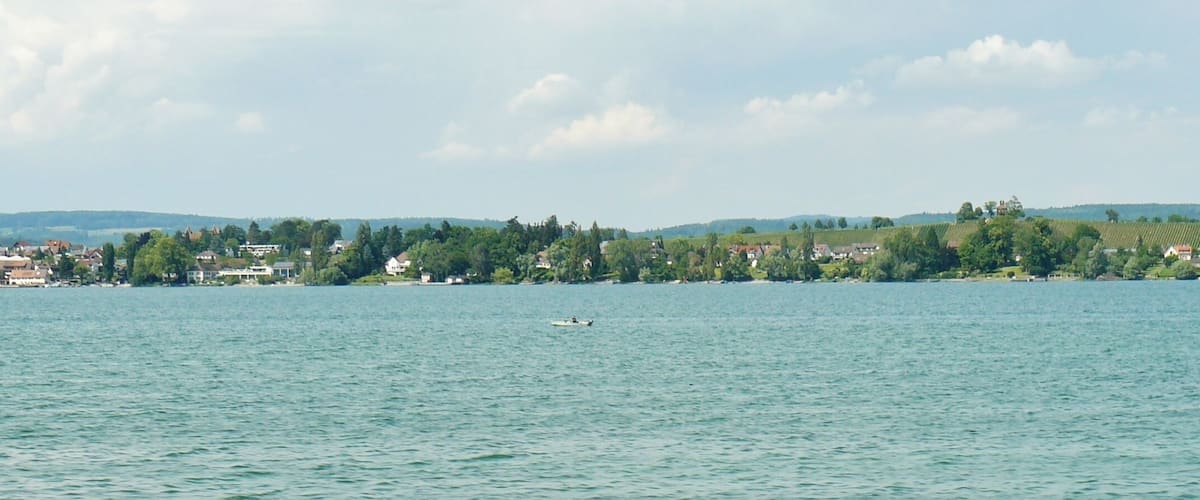 A calm lake with a small boat and a buoy. In the background, there is a town with houses and trees along the shore.