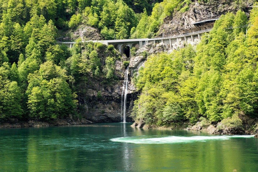 Un pont enjambant une rivière avec une cascade, le tout entouré d’une forêt verdoyante et dense.