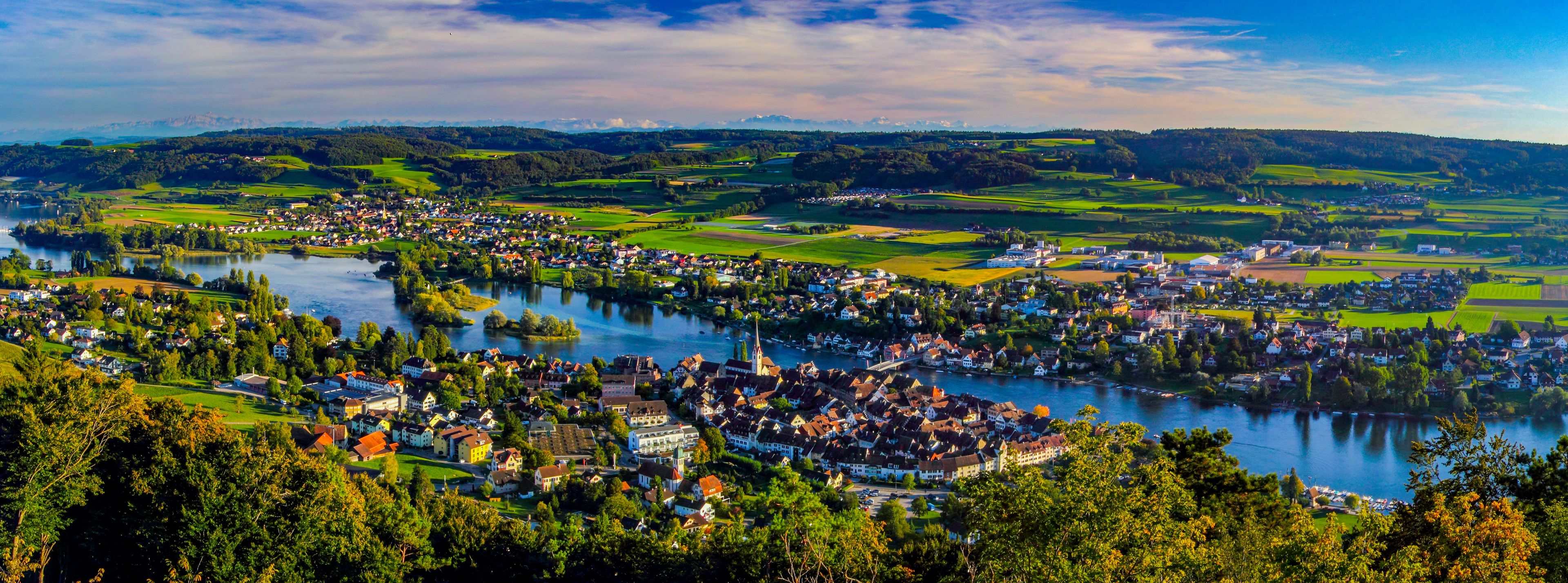 A river flowing through a town with surrounding green fields and mountains in the distance.