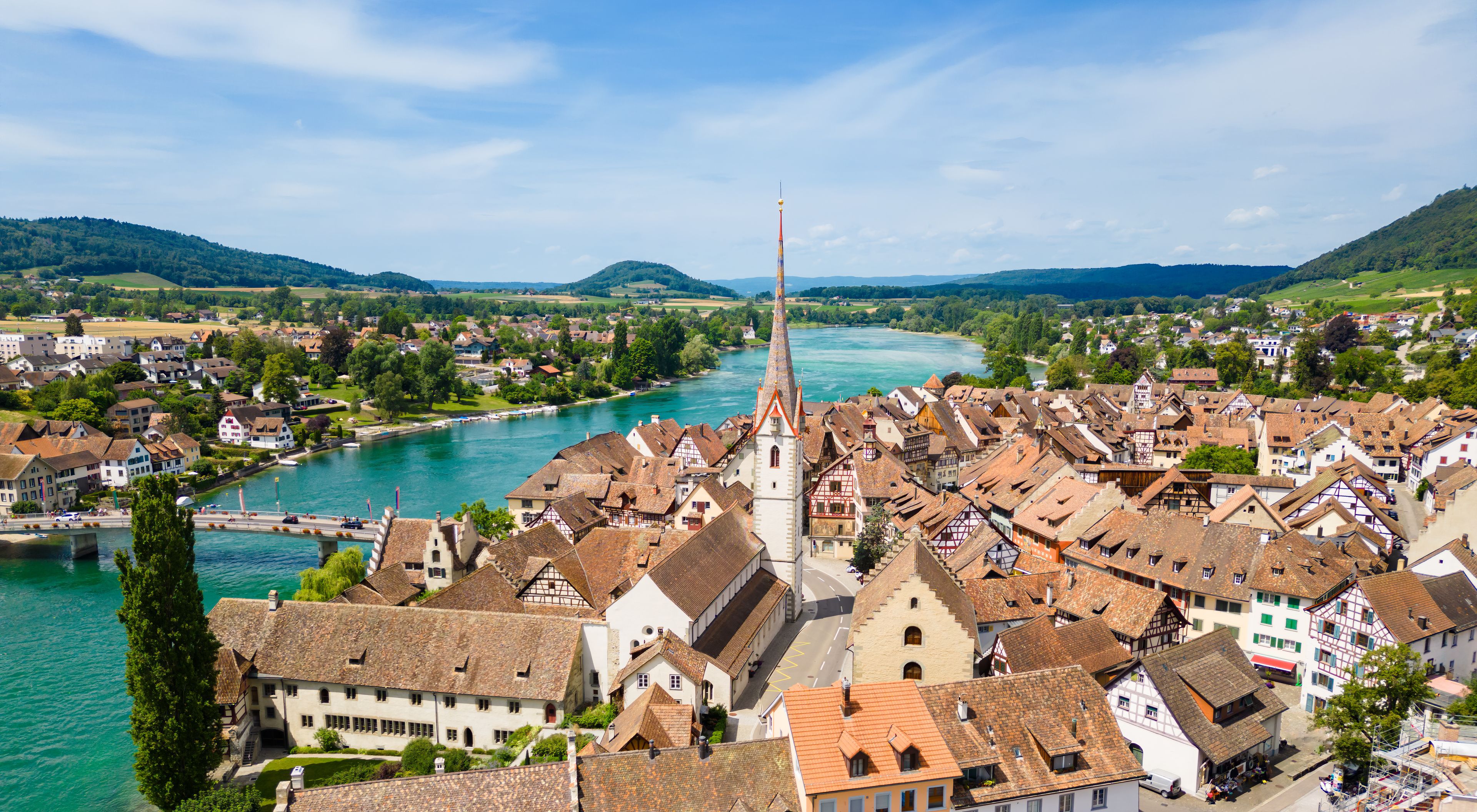 A riverside town with a prominent church steeple, traditional half-timbered houses, and a bridge connecting the town to the river.