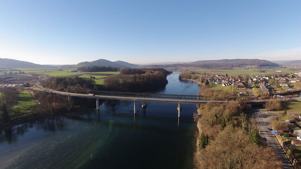 A river flowing through a valley with a bridge crossing over it, surrounded by fields and a small village.