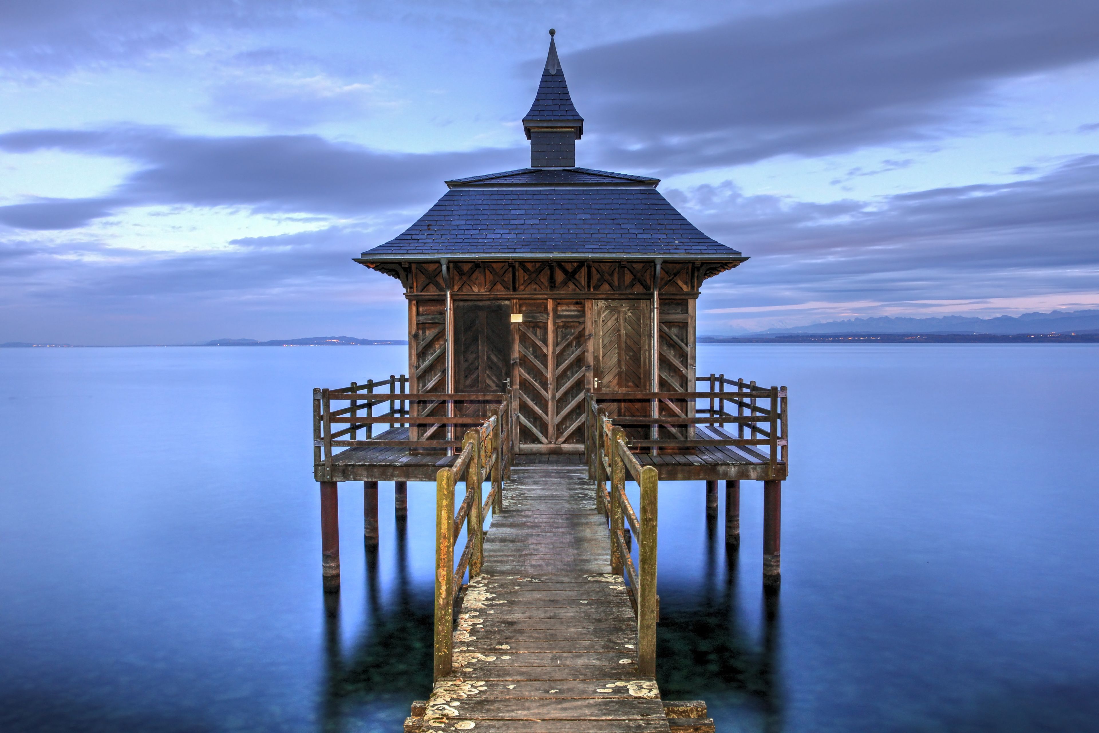 A wooden pier leading to a small, dark-roofed structure with a pointed roof, extending into a calm body of water with a distant shoreline.
