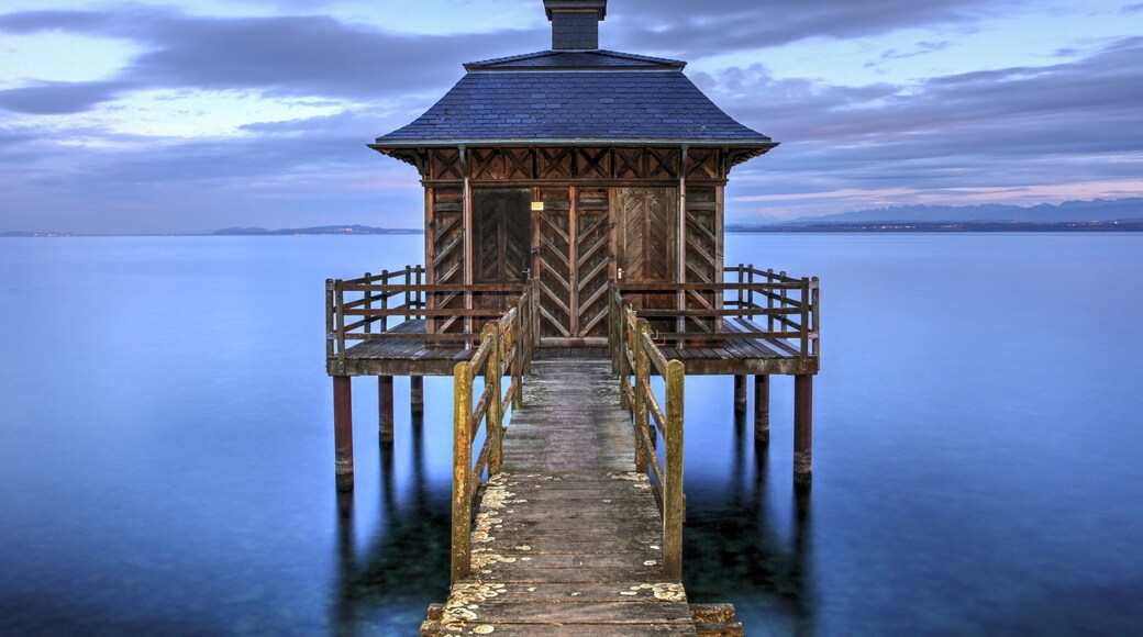 A wooden pier leading to a small, dark-roofed structure with a pointed roof, extending into a calm body of water with a distant shoreline.