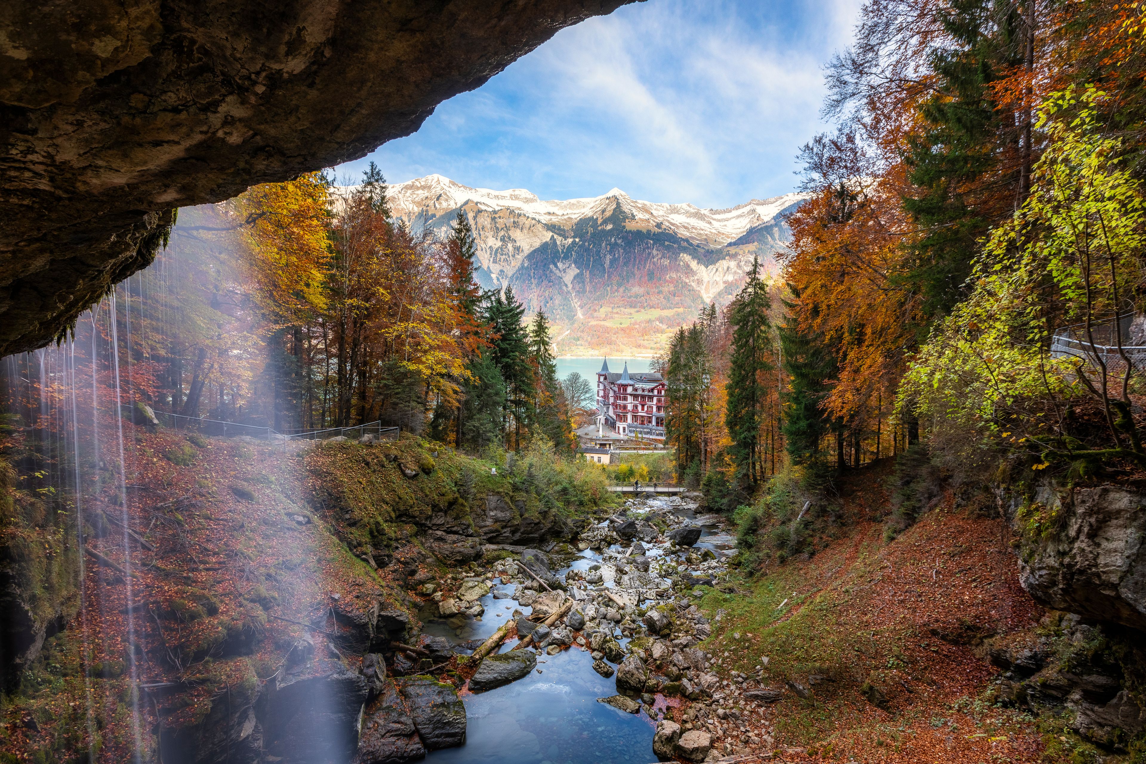 Une rivière traverse une forêt où les arbres arborent les couleurs de l’automne, une chaîne de montagnes se profile à l’arrière-plan, et un bâtiment doté d’un toit rouge se dresse.