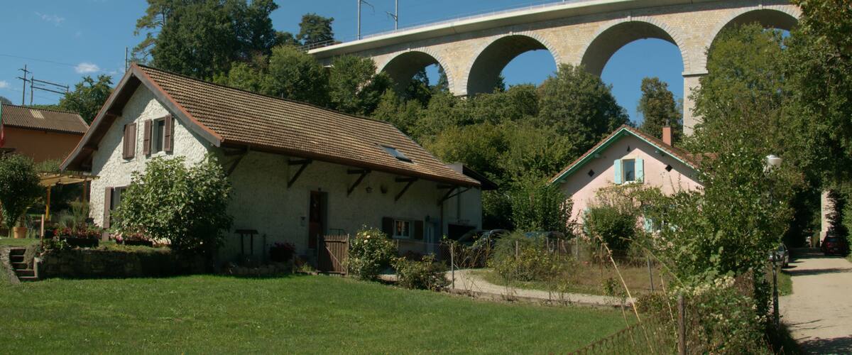 A house with a tiled roof, a stone bridge with arches, and lush green trees.