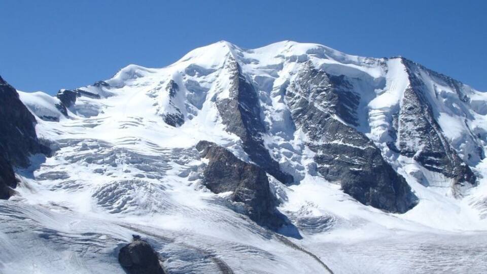 A snow-covered mountain with rocky terrain and two people standing at the base.