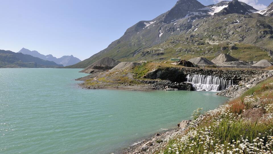 A mountainous landscape with a waterfall, a lake, and a rocky shoreline with wildflowers.