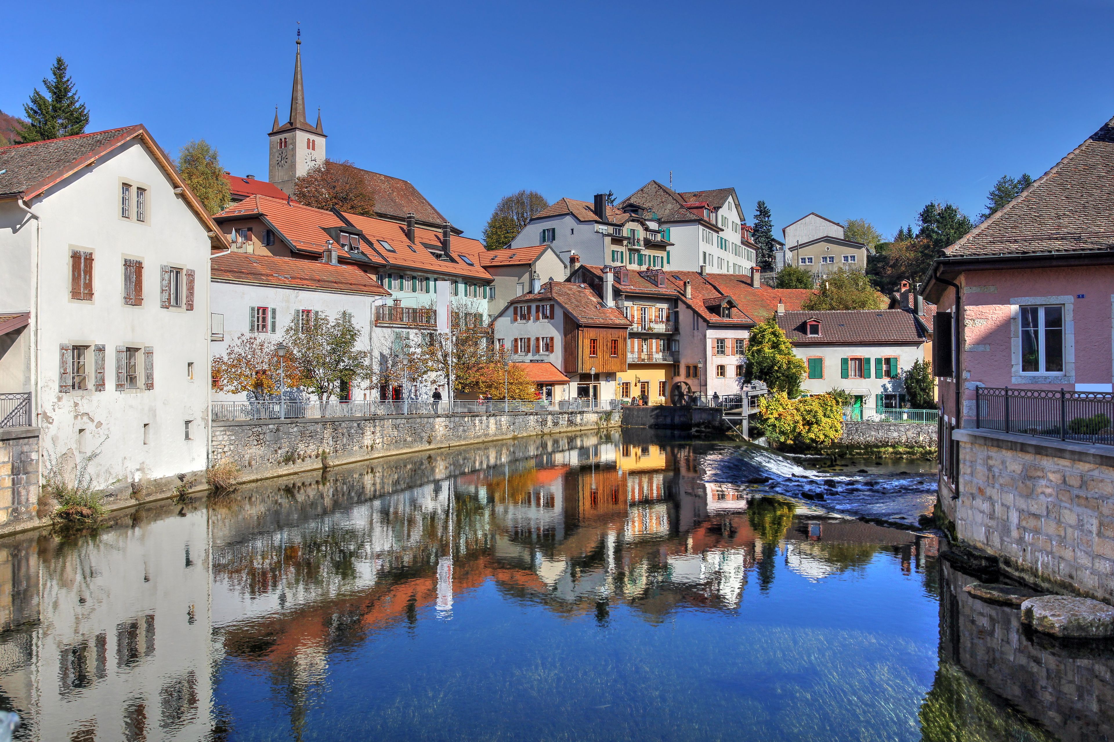A calm river reflecting buildings, a church steeple, and autumn trees.