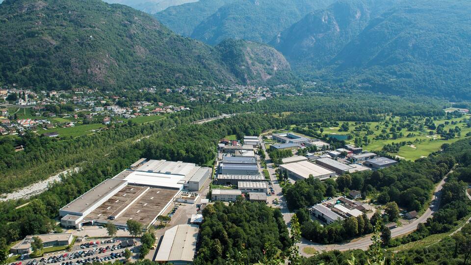 An industrial area with large buildings and a parking lot, surrounded by lush green hills and mountains.