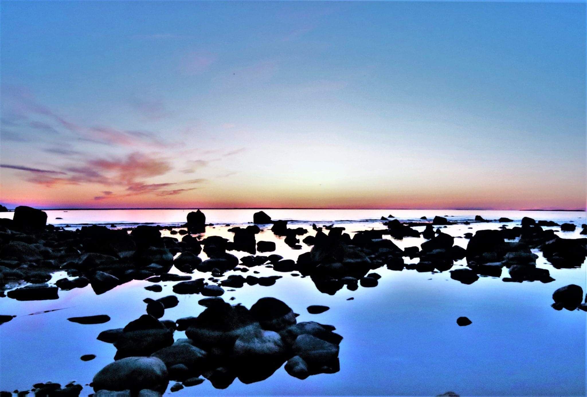 A rocky shoreline at sunset with calm water reflecting the sky.