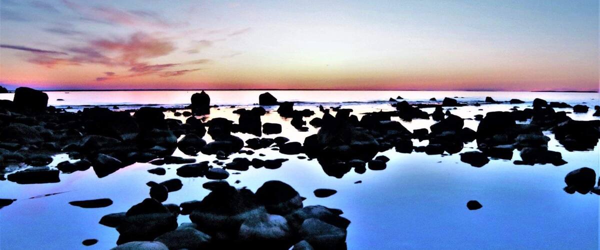 A rocky shoreline at sunset with calm water reflecting the sky.