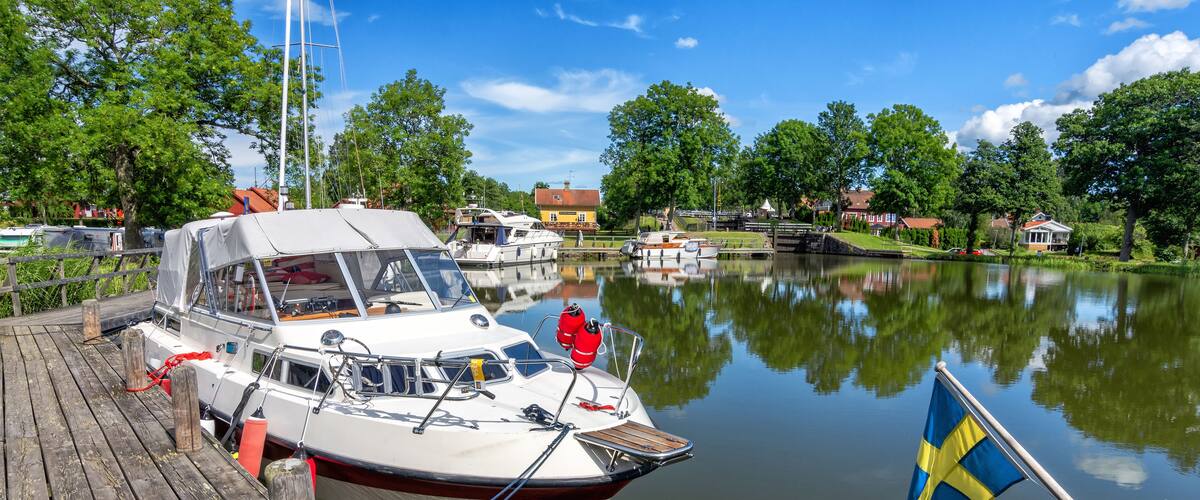 A calm lake with a wooden dock, a boat with a Swedish flag, and houses in the background.