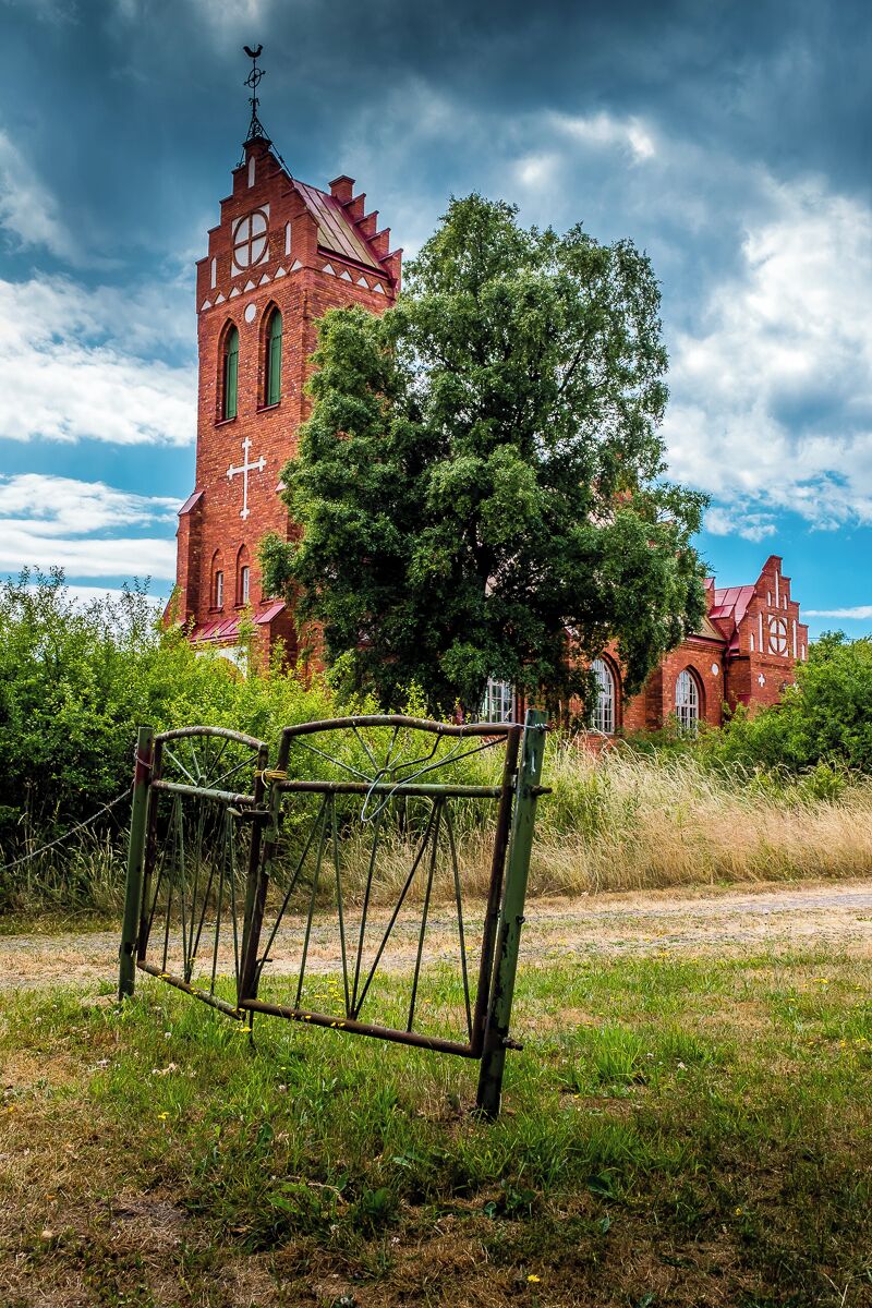 A red brick church with a tall tower and a cross, surrounded by trees and a metal gate.