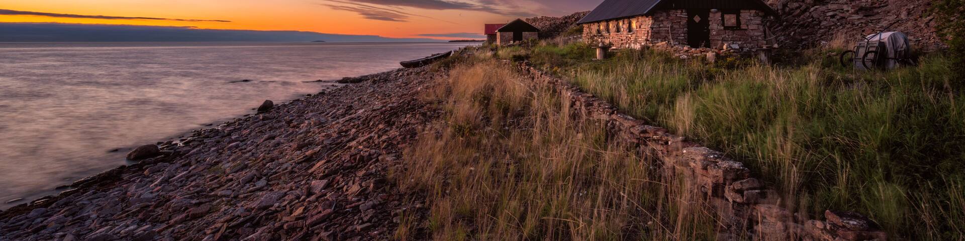 A stone house by a rocky shoreline with a sunset in the background.