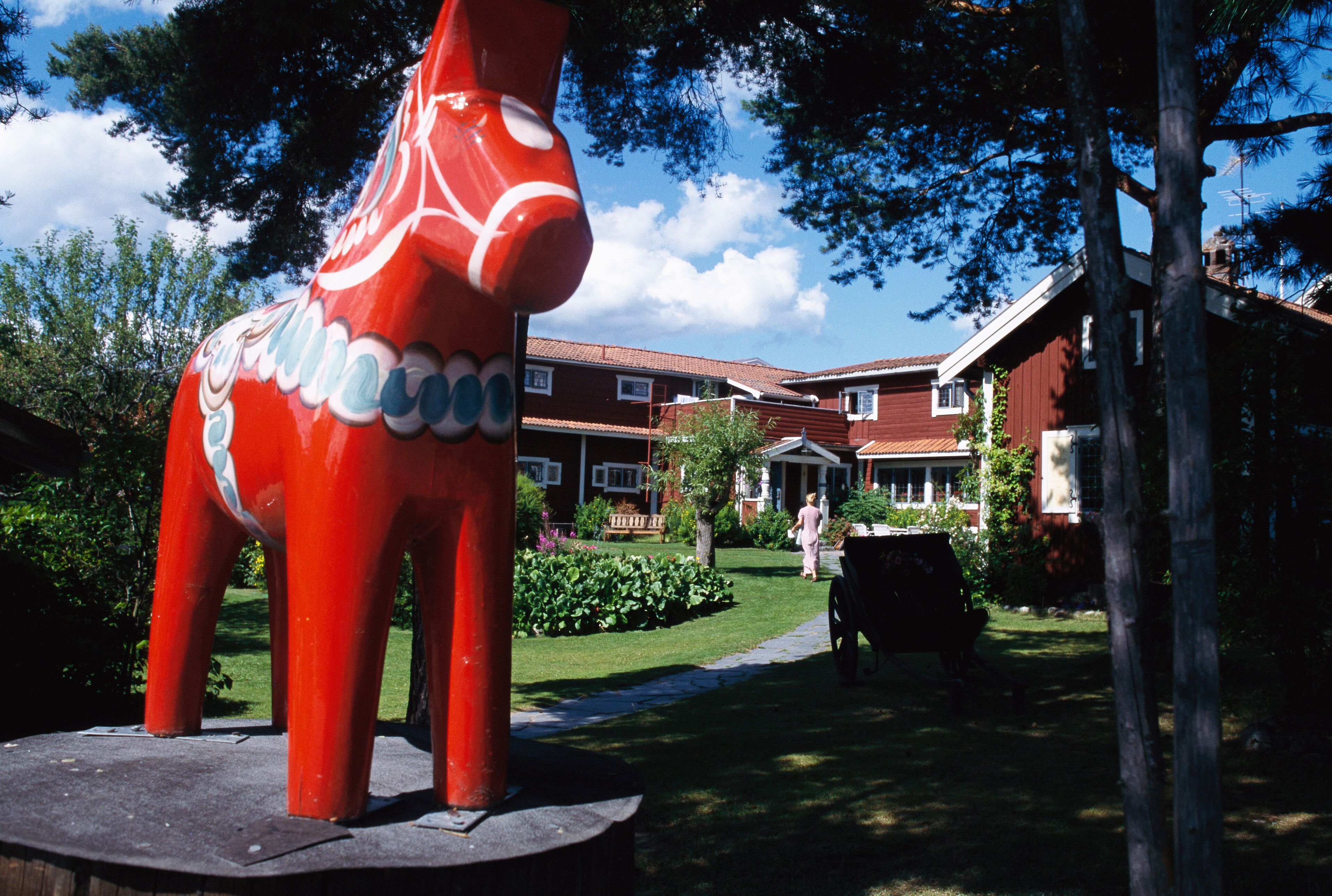 A red horse sculpture in a garden with a house and trees in the background.