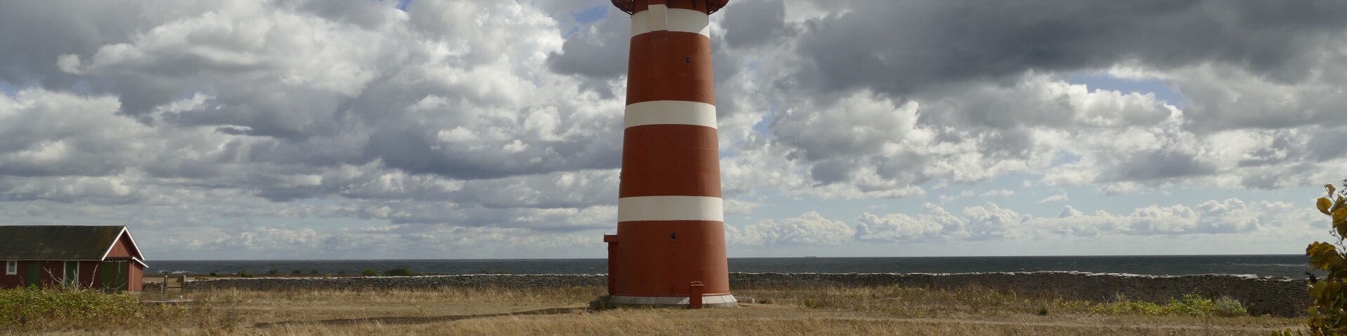 A red and white striped lighthouse with a green roof, standing on a grassy field with a stone wall in the foreground.