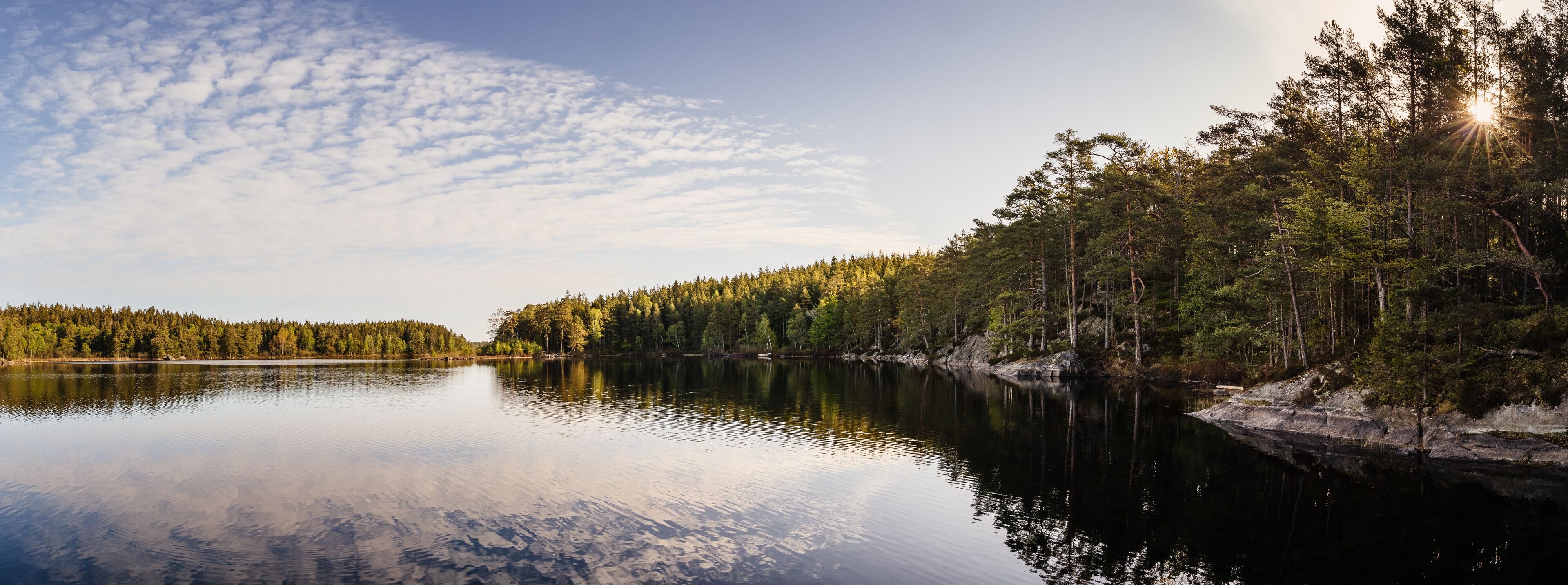 A calm lake surrounded by dense forest and a clear sky with scattered clouds.