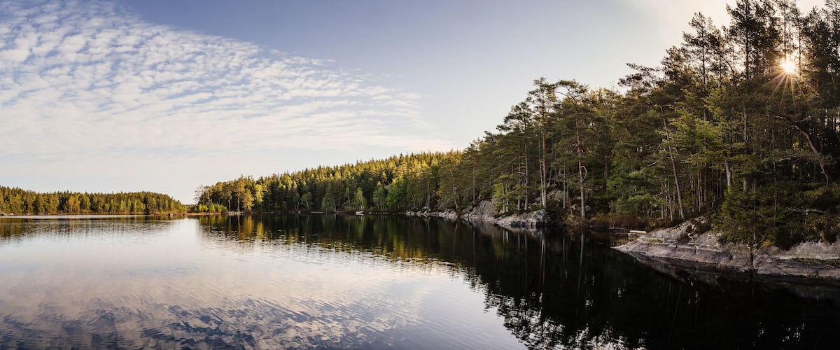 A calm lake surrounded by dense forest and a clear sky with scattered clouds.
