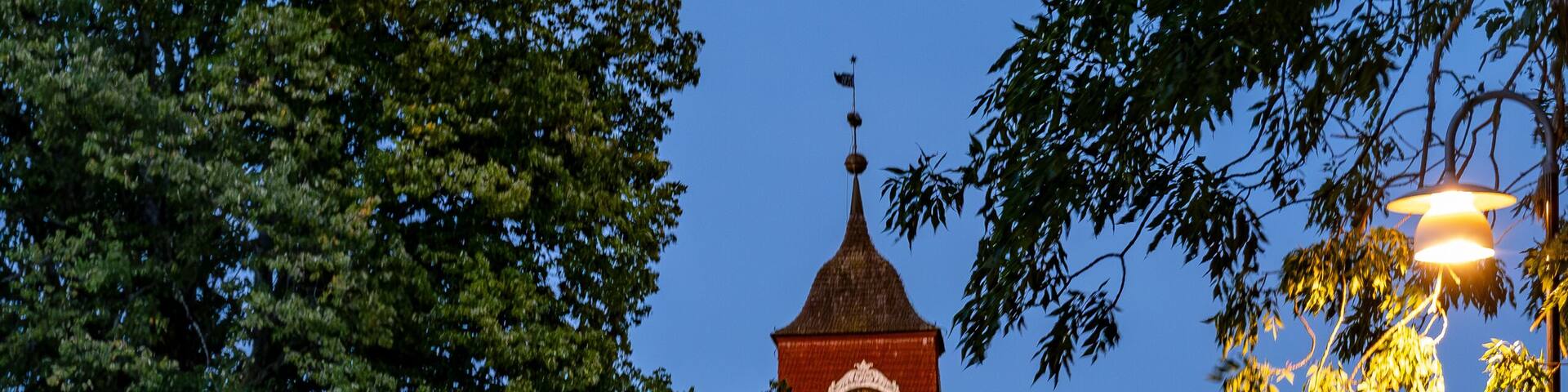 A red clock tower with a pointed roof, surrounded by trees and a road.