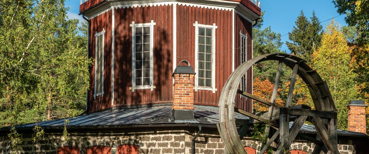 A historic building with a large wooden water wheel, surrounded by trees and greenery.