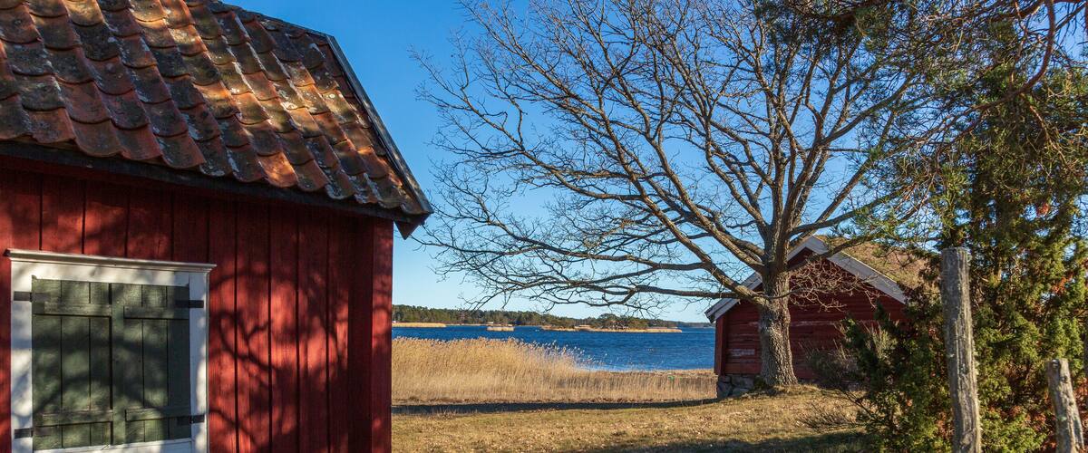 A red wooden building with a tiled roof, a tree, and a body of water in the background.