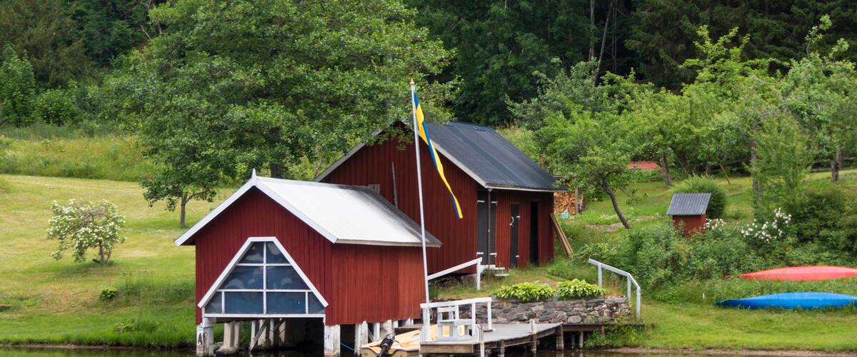 A red boathouse on a calm lake with a flagpole, surrounded by lush greenery and a small dock.