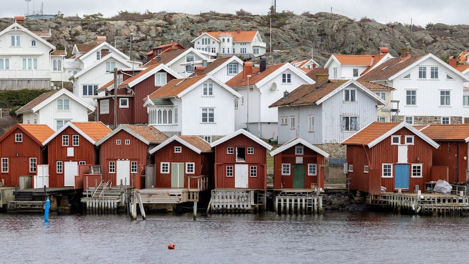 A cluster of wooden houses with red roofs and white walls, situated by a body of water with rocky hills in the background.