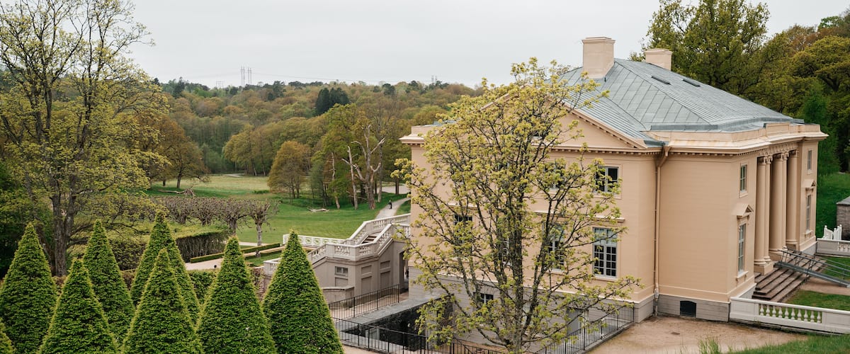 A large, beige building with a green roof, surrounded by well-manicured trees and a grassy area.