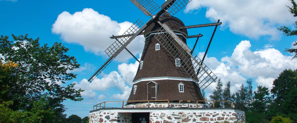 A traditional windmill with a stone base, surrounded by greenery and a clear blue sky.