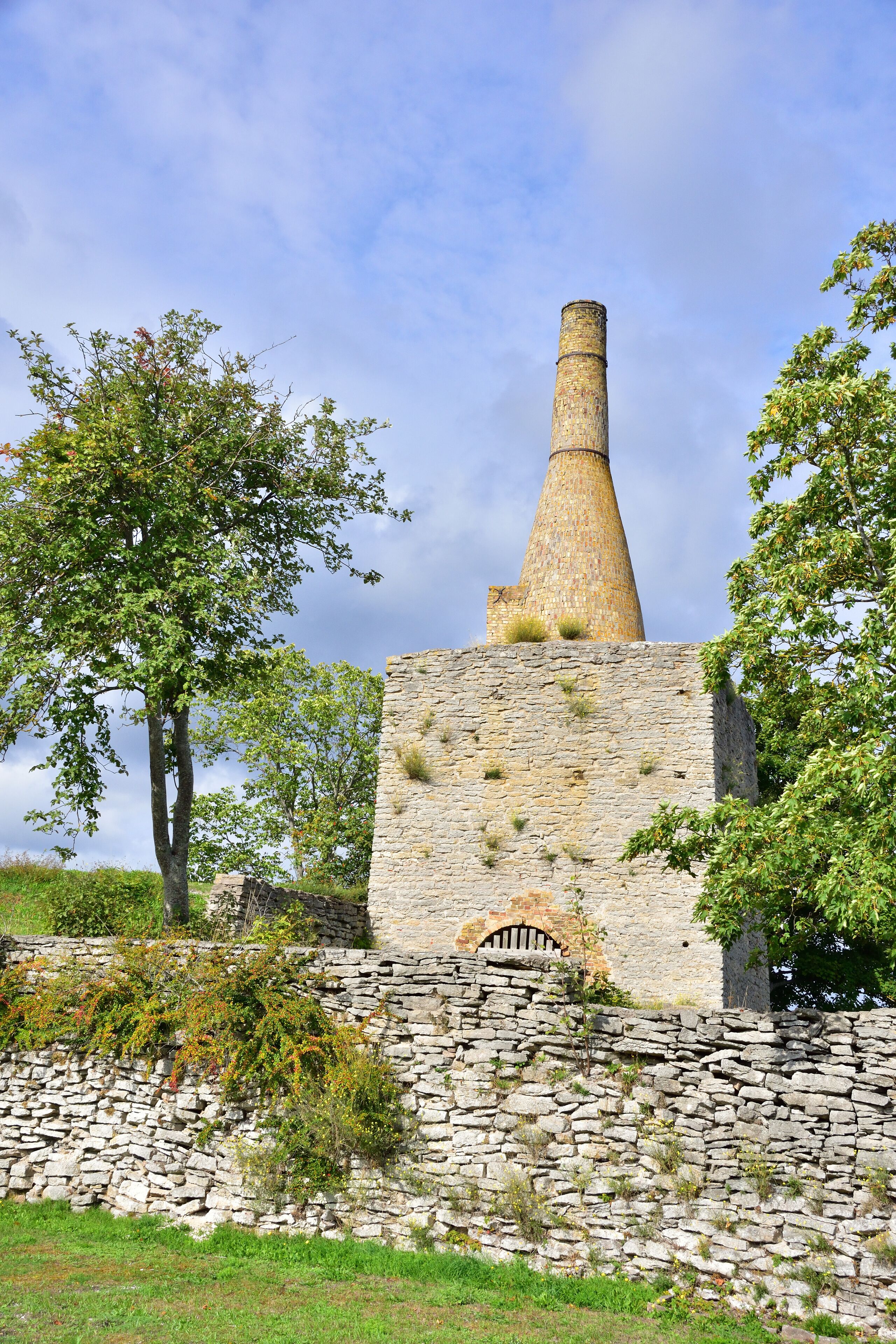 A stone structure with a conical chimney and a stone wall with vegetation.