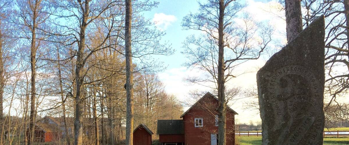 A rural landscape with a red barn, a small house, and a stone monument in a grassy field with trees and a stream.