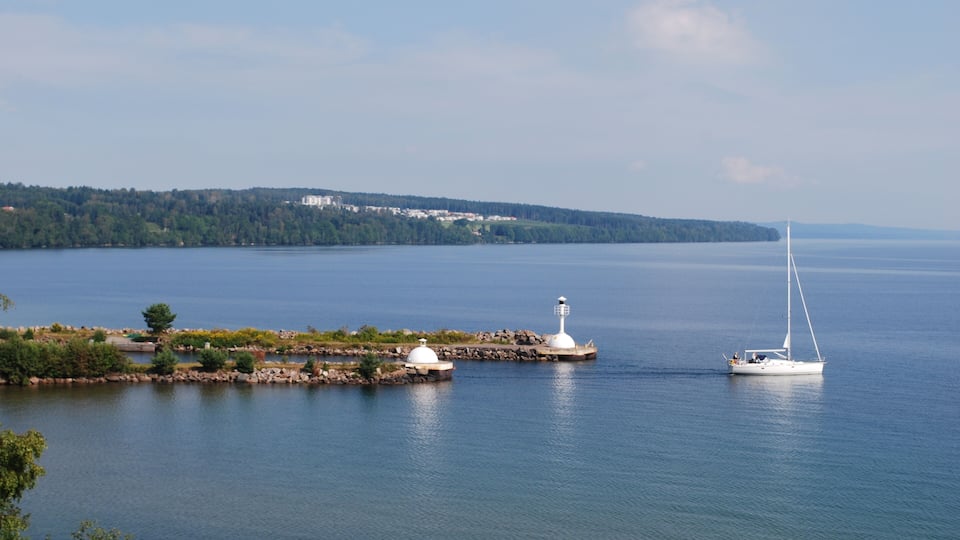 A calm lake with a sailboat, a lighthouse, and a small island with buildings in the background.