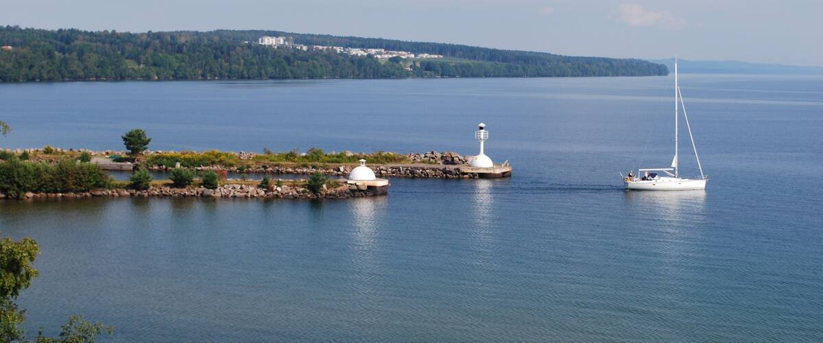 A calm lake with a sailboat, a lighthouse, and a small island with buildings in the background.