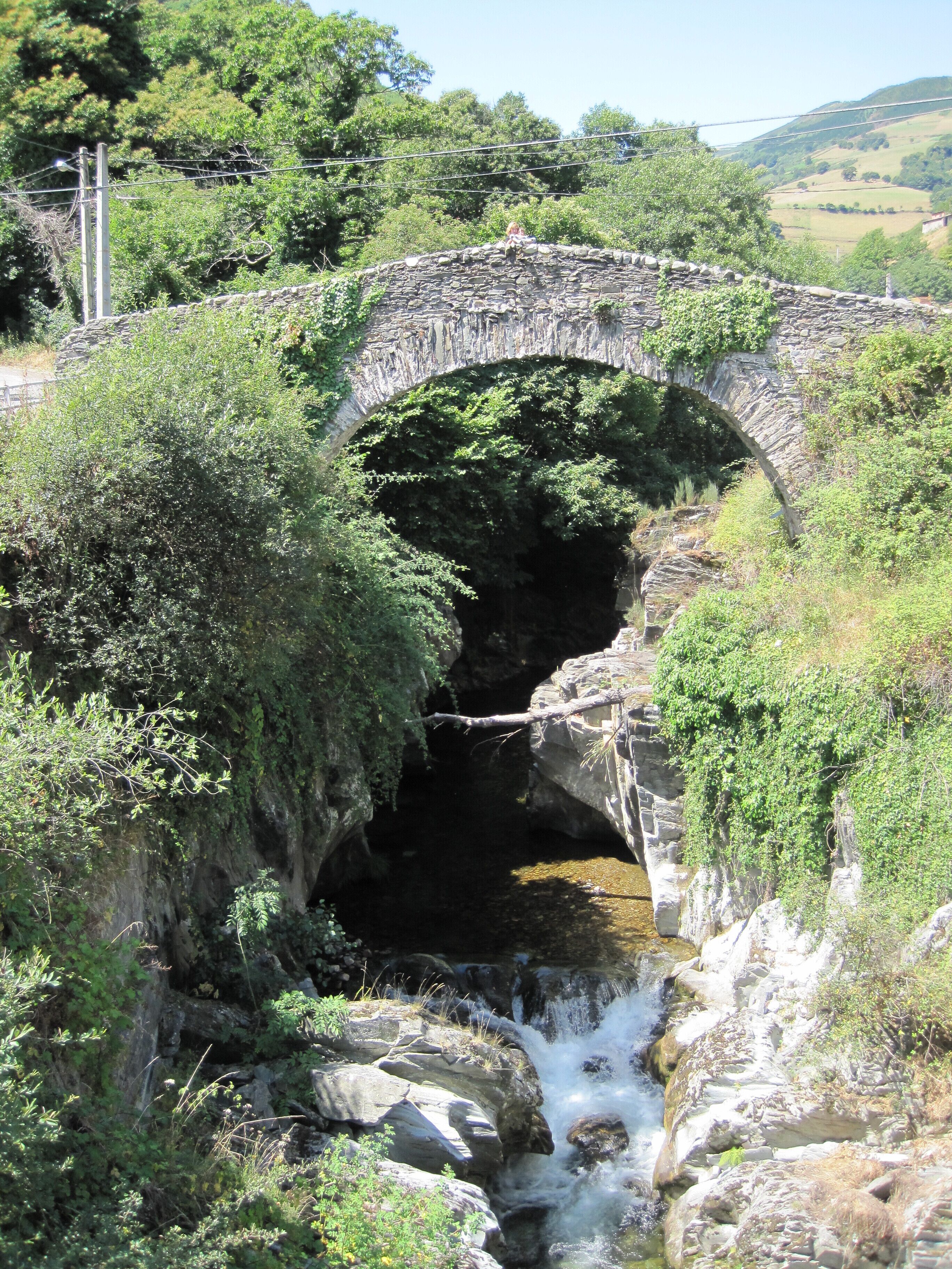 A stone bridge over a stream with lush greenery and hills in the background.