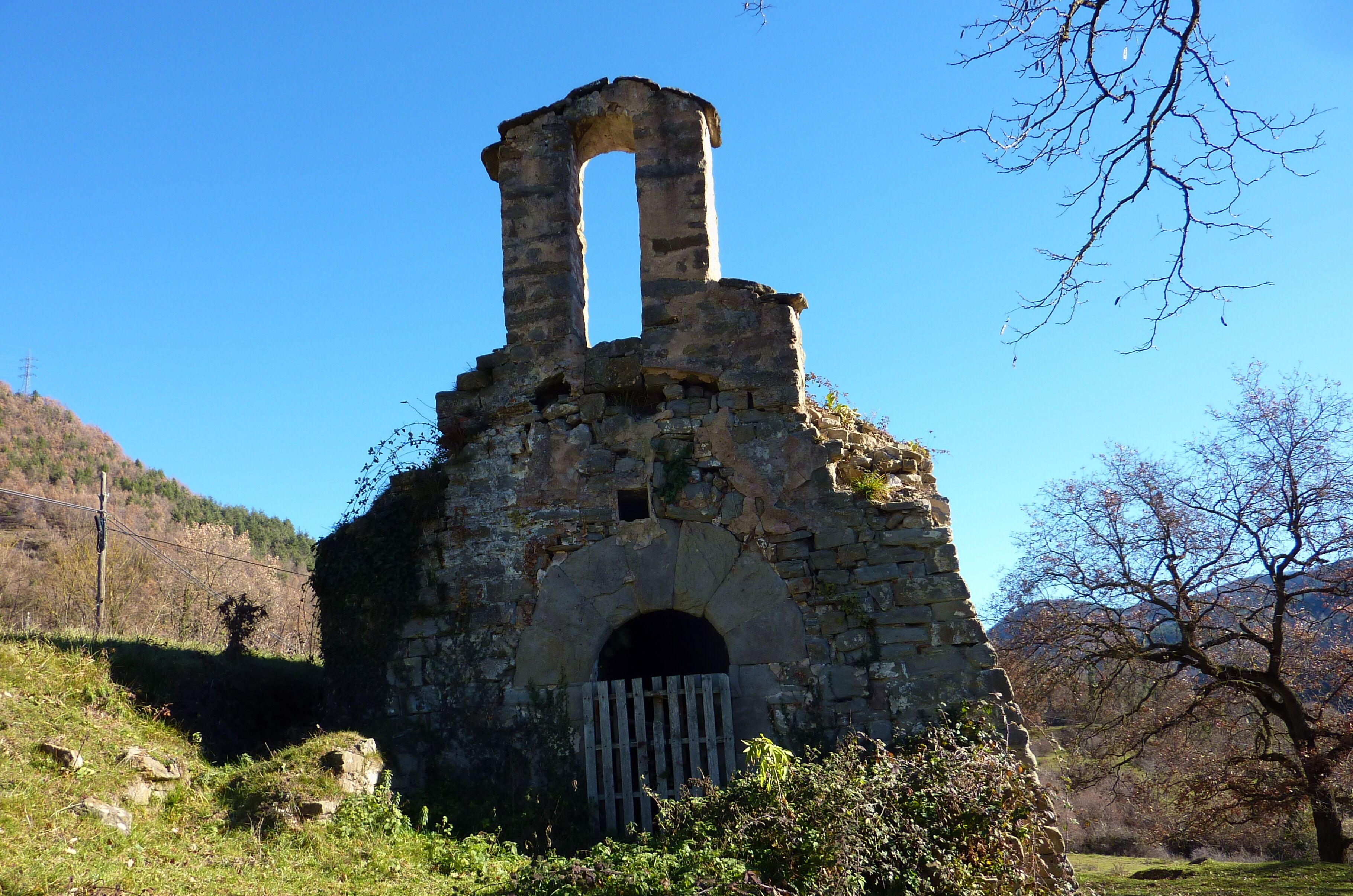 An ancient stone ruin with a central archway and a tall, narrow tower, surrounded by trees and hills.