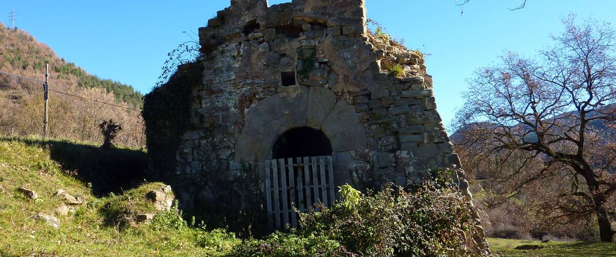 An ancient stone ruin with a central archway and a tall, narrow tower, surrounded by trees and hills.