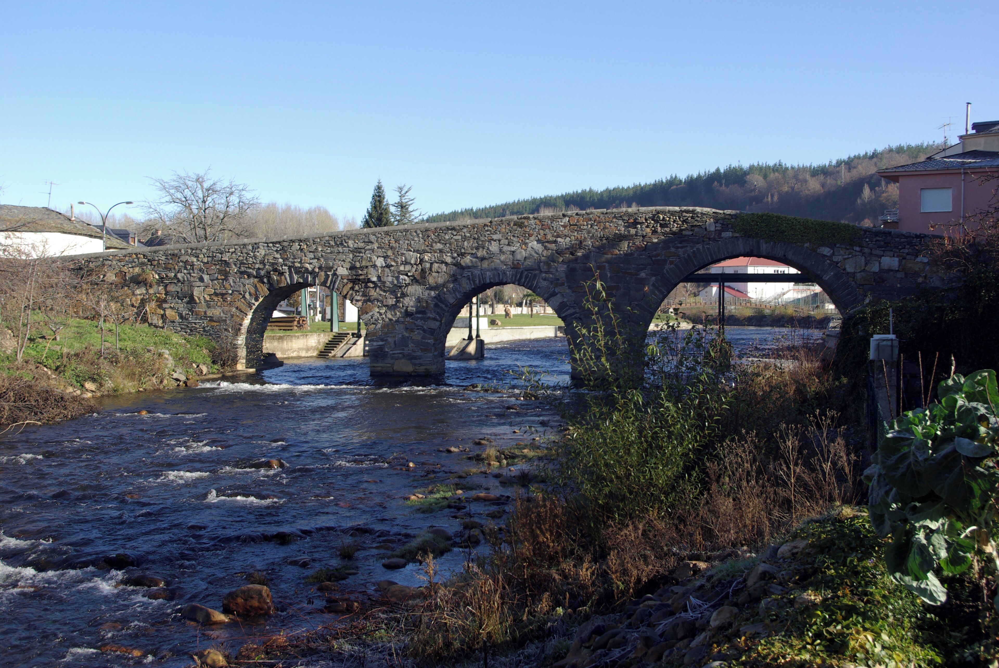 A stone bridge over a flowing river with a clear sky and surrounding greenery.