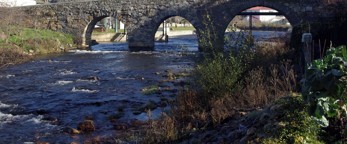 A stone bridge over a flowing river with a clear sky and surrounding greenery.